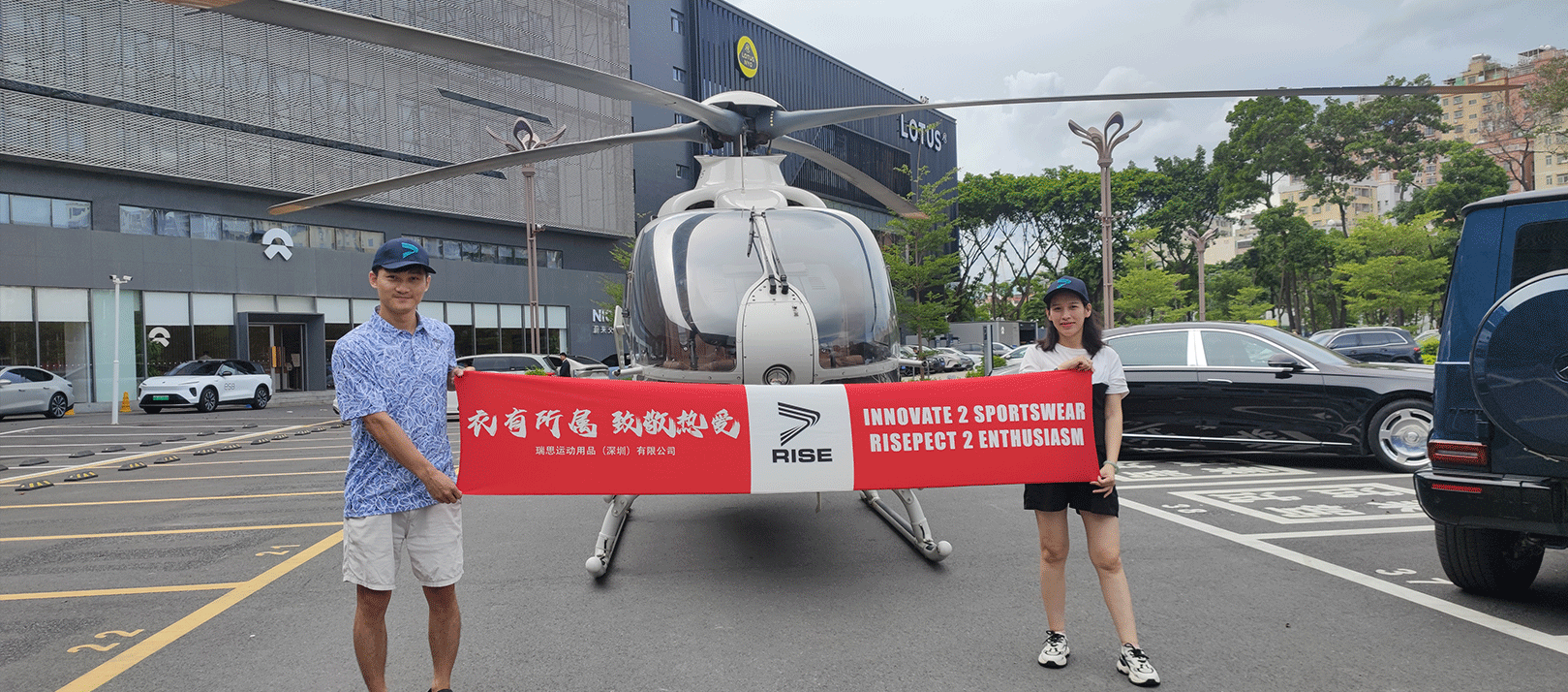 The boss decked out in Rise golf polo shirt, and his wife, taking pictures in front of the helicopter while holding a red banner that reads Clothes belong to someone, Hats off ardently love and Innovate 2 sportswear, risepect 2 enthusiasm